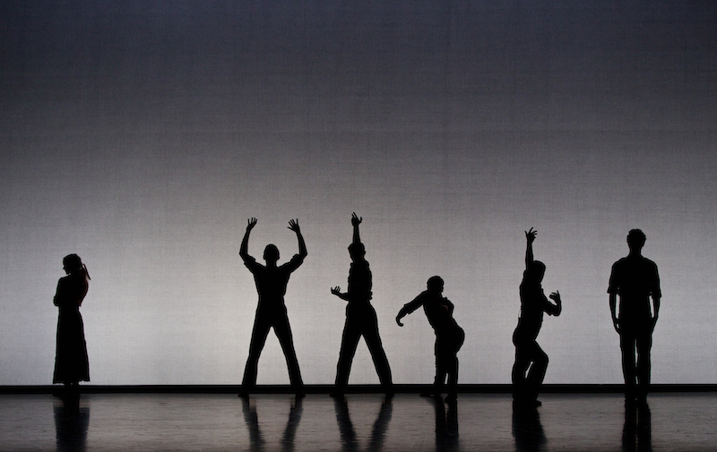 A group of dancers in silhouette. They're positioned at varying heights and assuming different poses. One figure is set apart with her arms crossed.